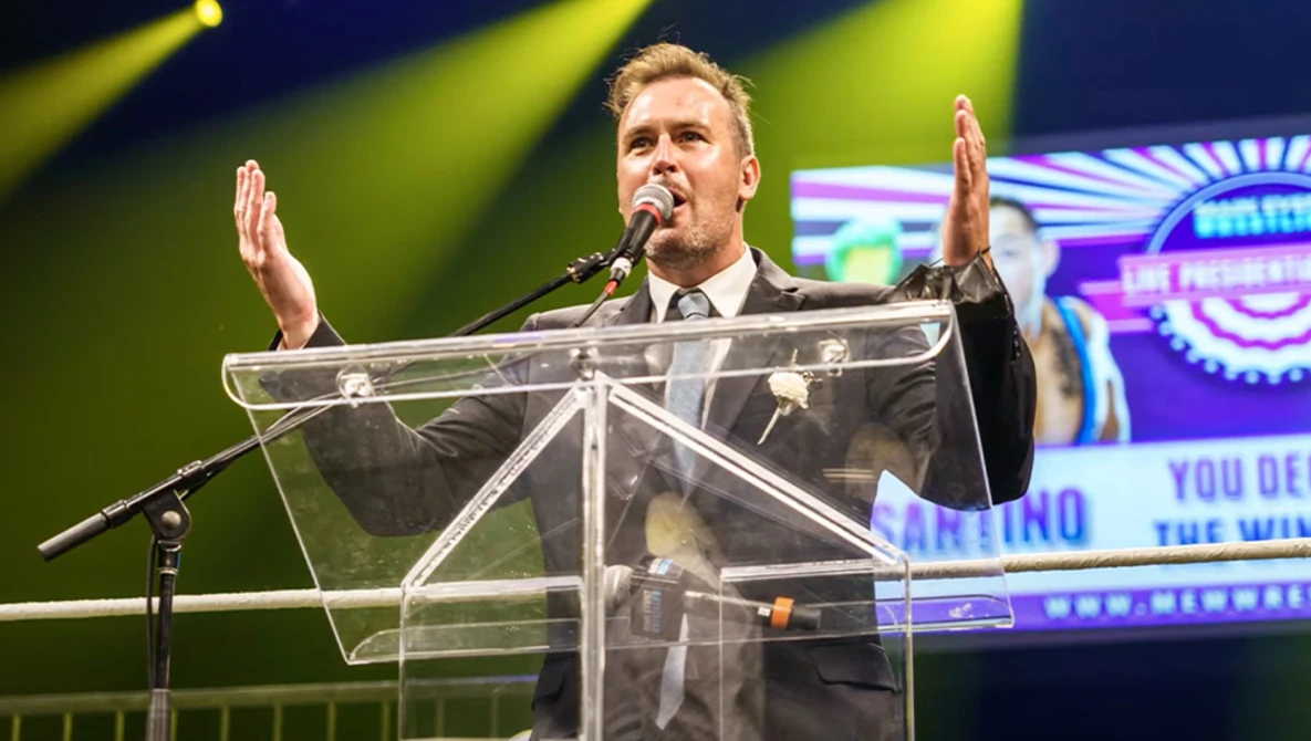 Man in business suit speaking at transparent podium with microphone, hands raised expressively.
