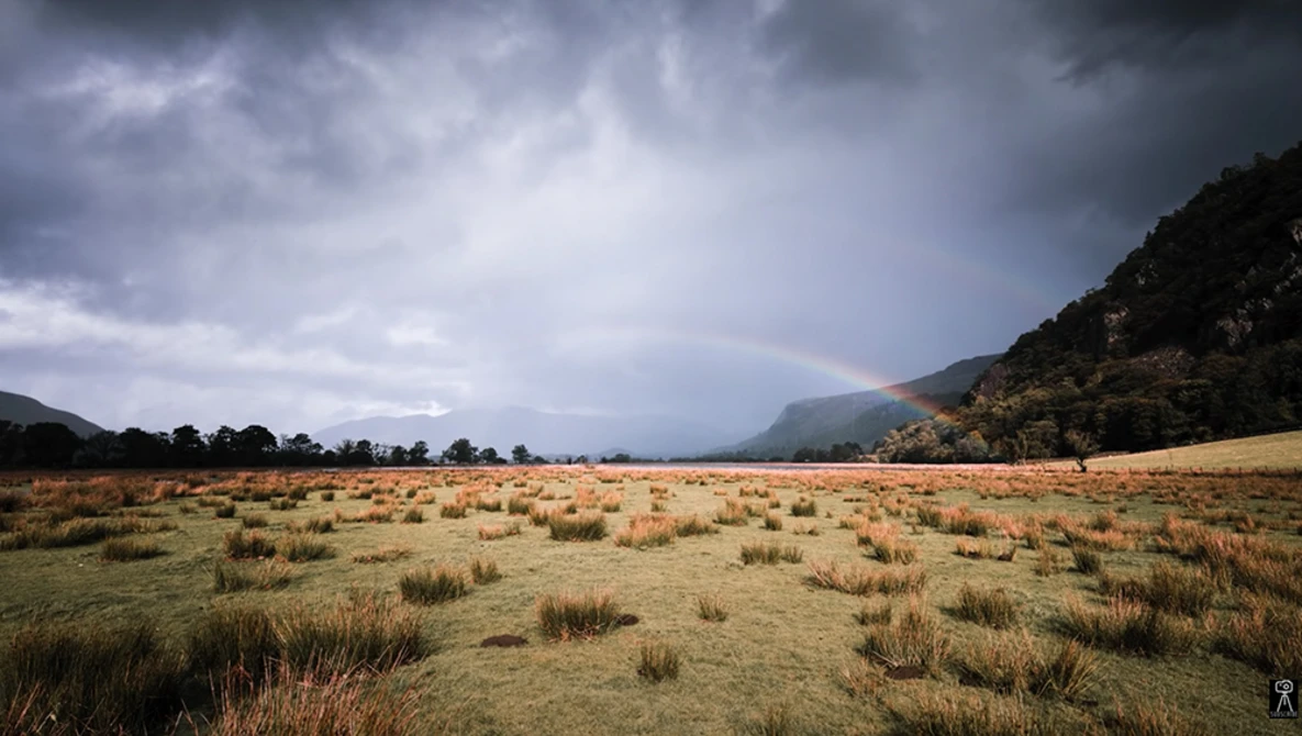 Expansive grassland valley beneath dramatic storm clouds, with forested mountains flanking both sides.