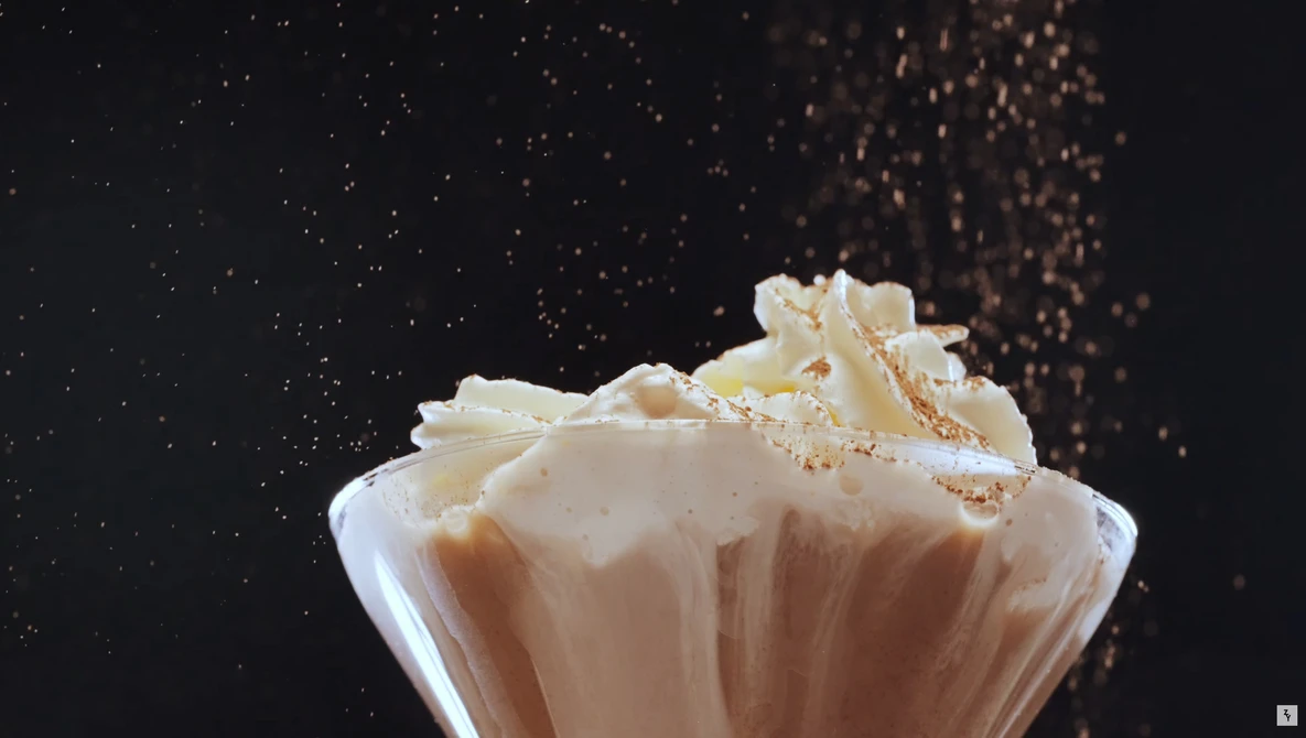 Creamy beverage in glass cup with splashing liquid and particles suspended in air against dark background.