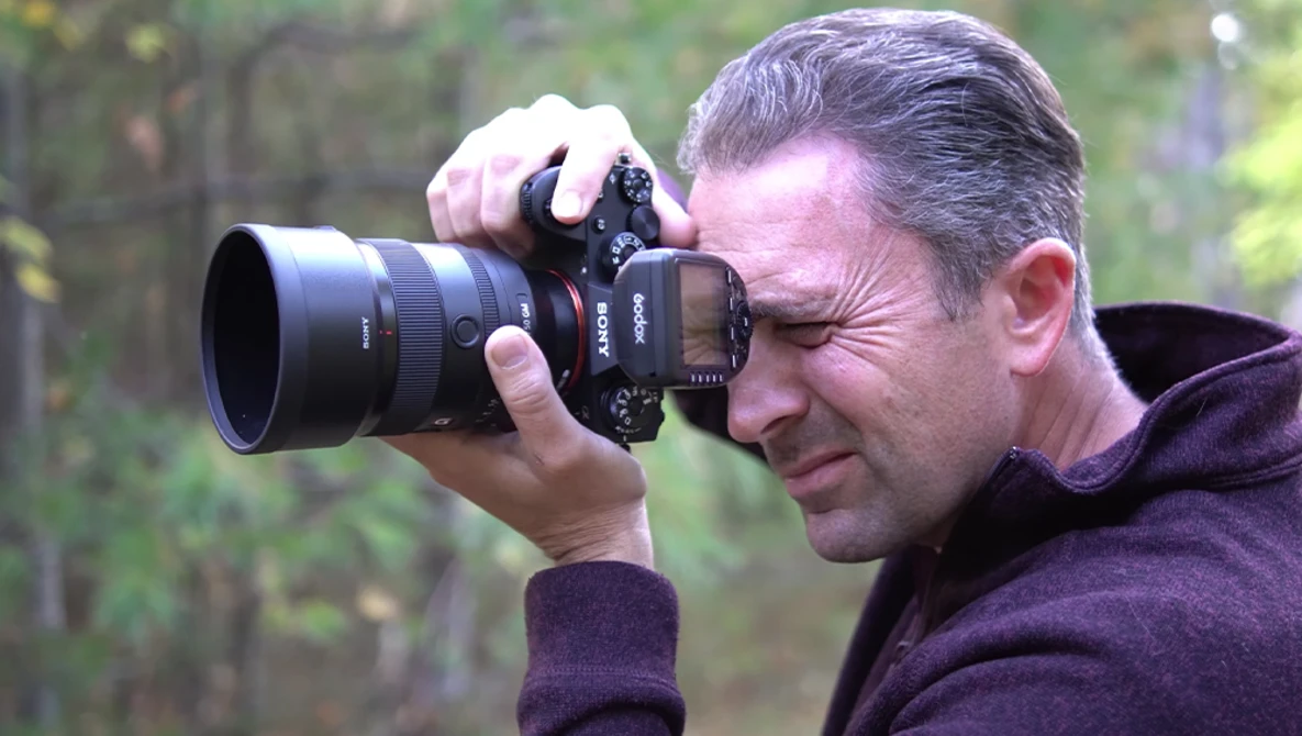 Male photographer holding a mirrorless camera with telephoto lens to his eye, focusing intently on a subject outdoors.