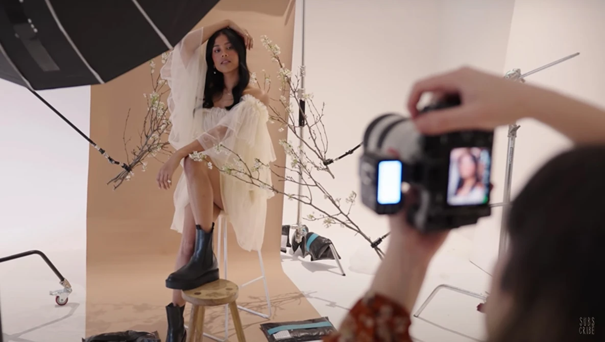 Behind-the-scenes view of a studio portrait shoot with model on stool and photographer operating camera in foreground.