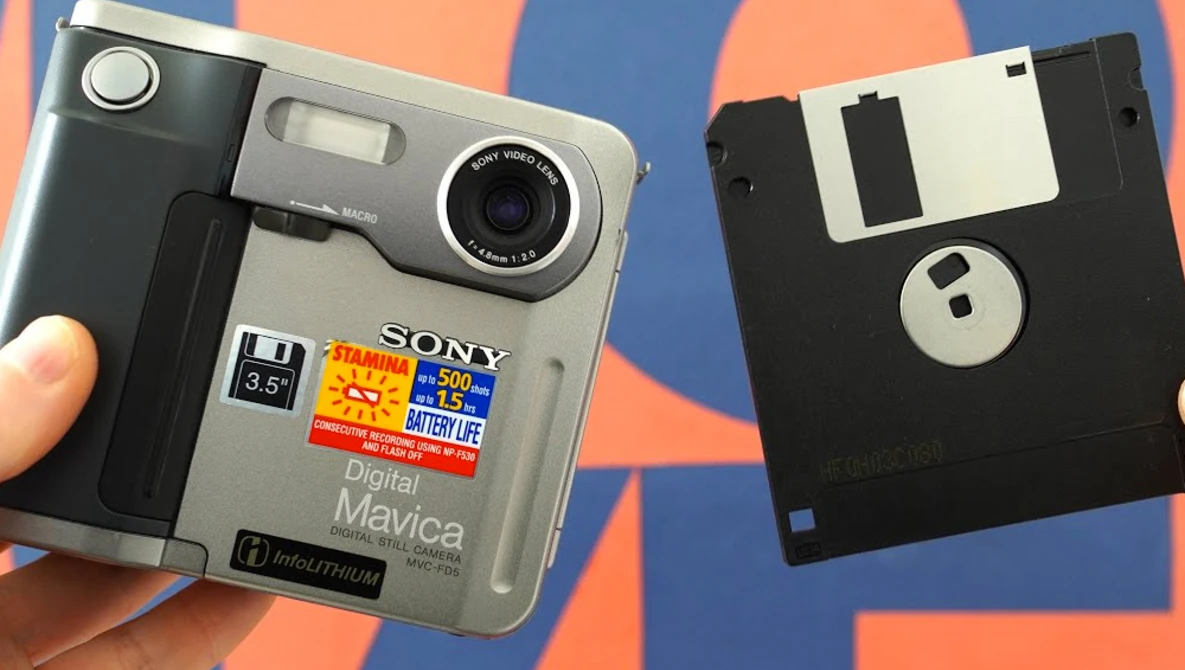 Sony Cyber-shot digital camera next to a floppy disk against a colorful striped background.