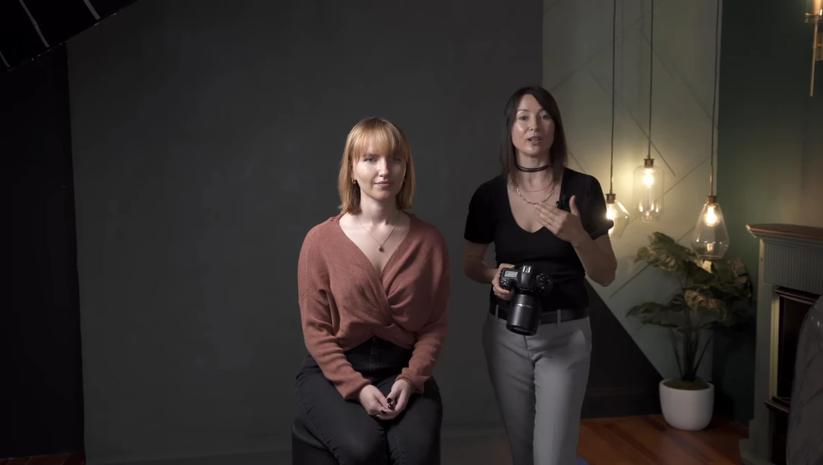 Two women in a modern indoor studio space with pendant lighting and plants in the background.