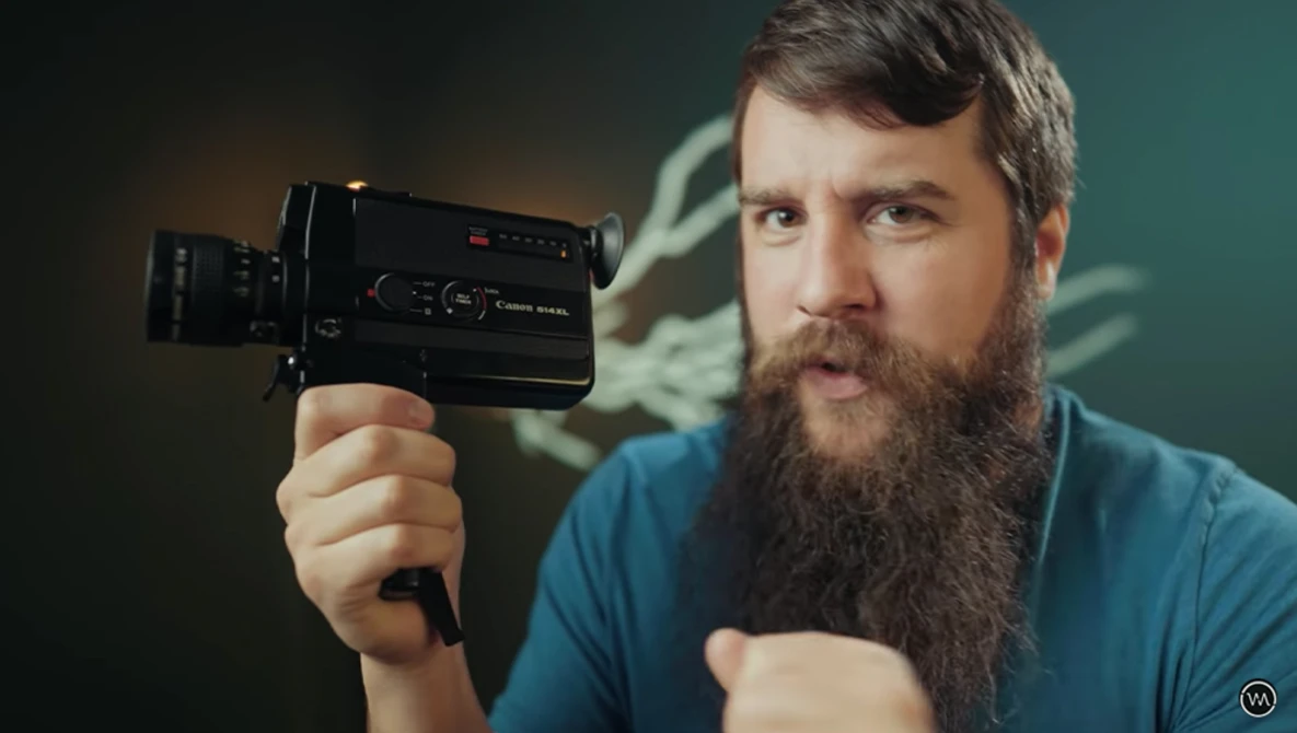 Bearded man in blue shirt holding a vintage film camera against a teal background.
