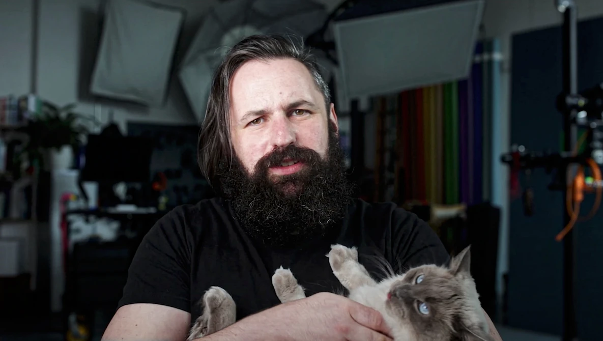 Bearded man holding a small tabby kitten in a studio setting with professional lighting equipment visible in background.