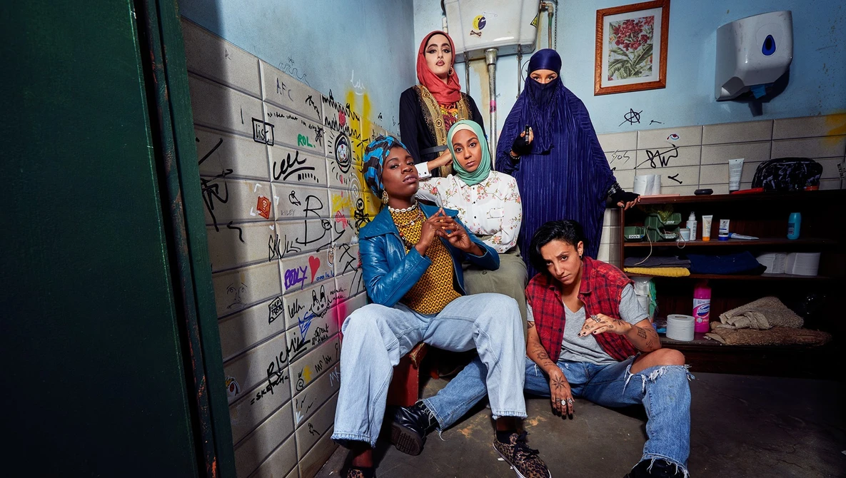 Six people of varying ages posing together in a modest room with blue walls and graffiti-covered surfaces.