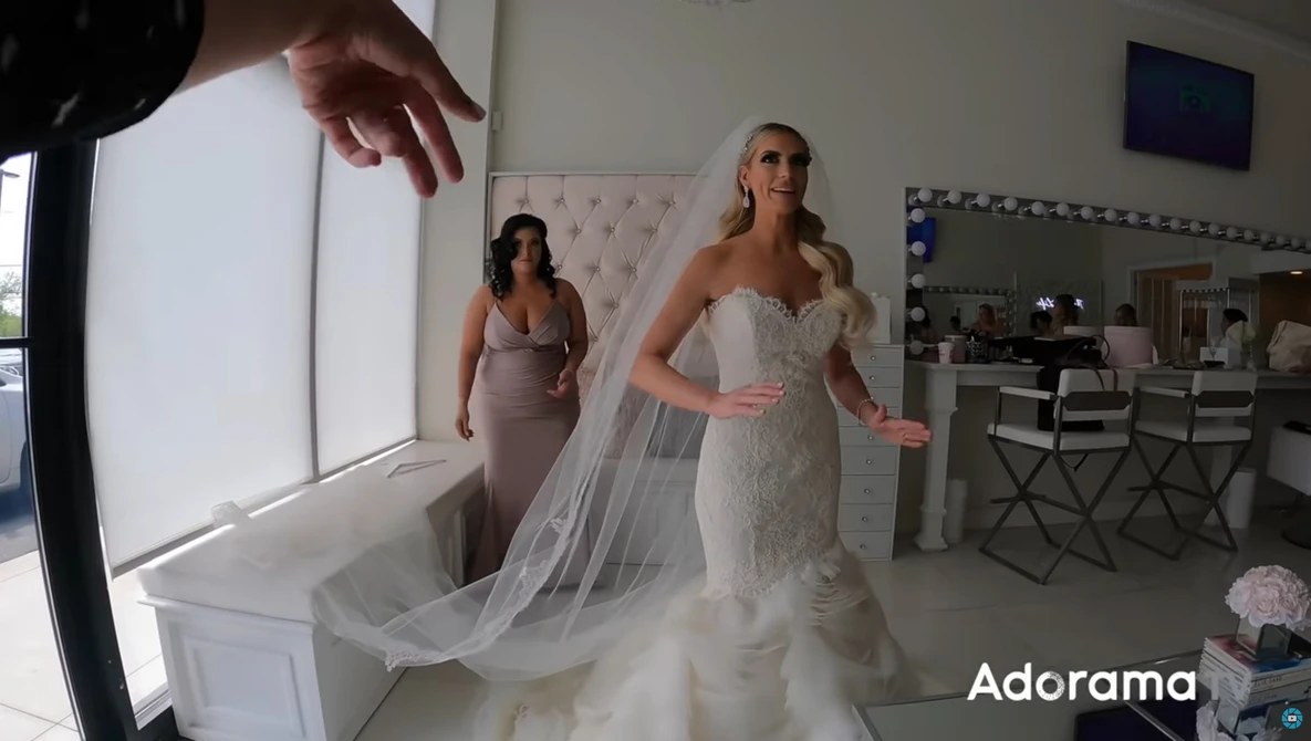 Bride in beaded wedding dress with flowing veil being adjusted during bridal preparation in salon.