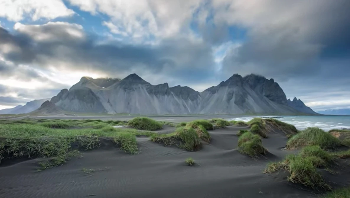 Dramatic mountain peaks rise behind a dark volcanic sand beach with sparse green vegetation and tidal pools.