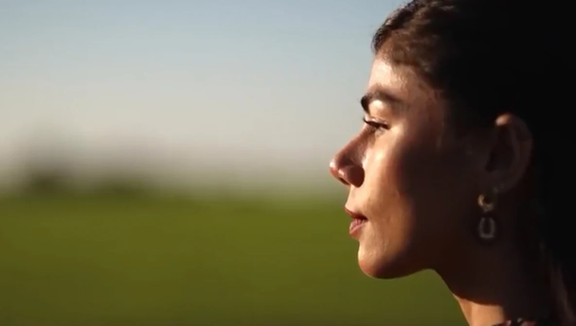 Woman in profile gazing toward a blurred green field under clear sky.