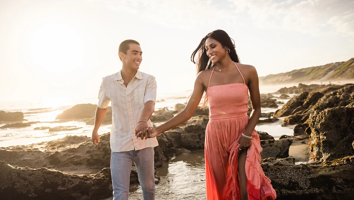 Couple holding hands walking along rocky coastline at golden hour sunset.