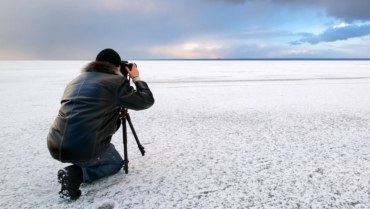 Photographer crouching on a frozen lake while composing a shot with a telephoto lens on a tripod.