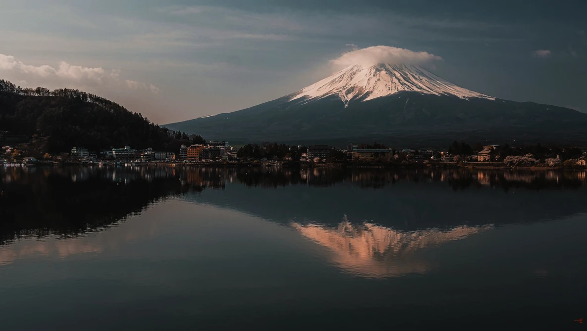 Snow-capped volcanic mountain reflected in still lake water at dawn, with town buildings along shoreline.