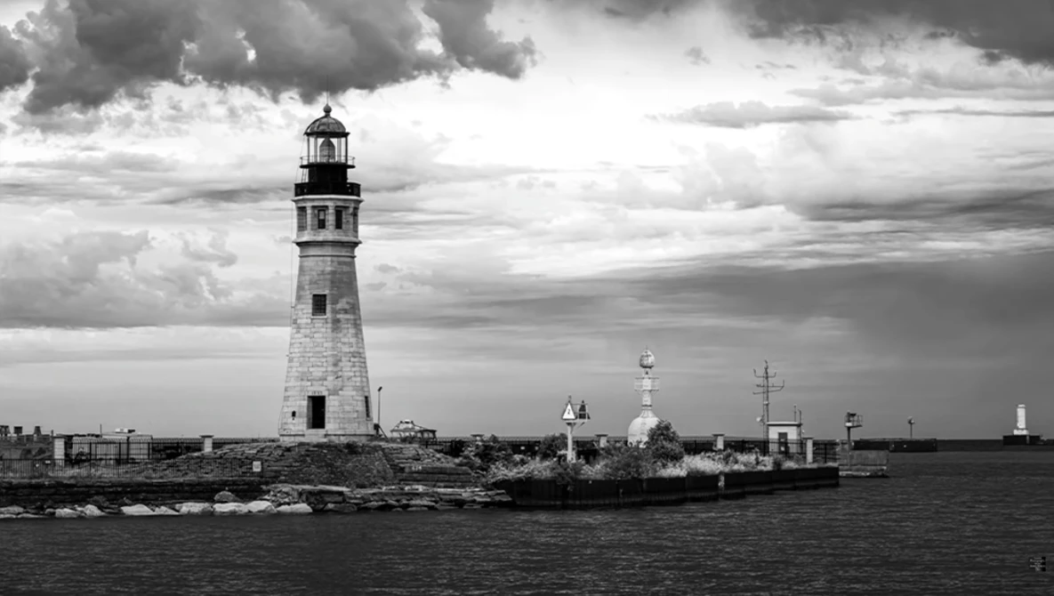 White lighthouse with lantern room stands on rocky shore, with industrial waterfront and dramatic cloudy sky in background.