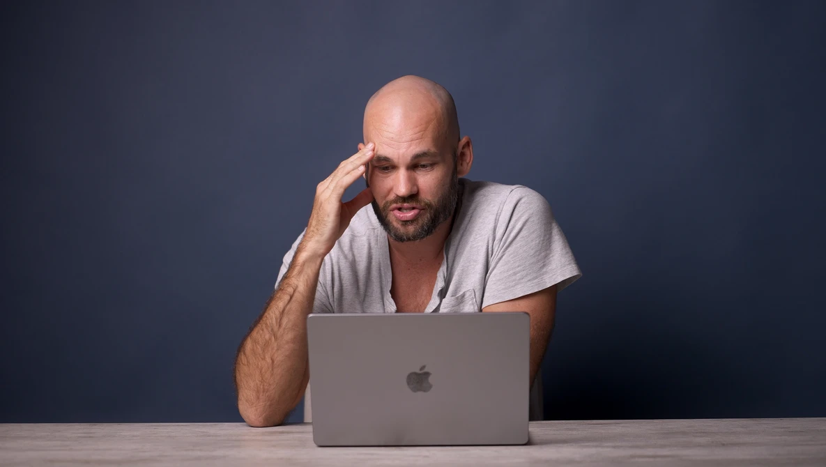 Bald man with beard wearing light gray v-neck shirt, leaning on desk with hand to face while looking at laptop screen.