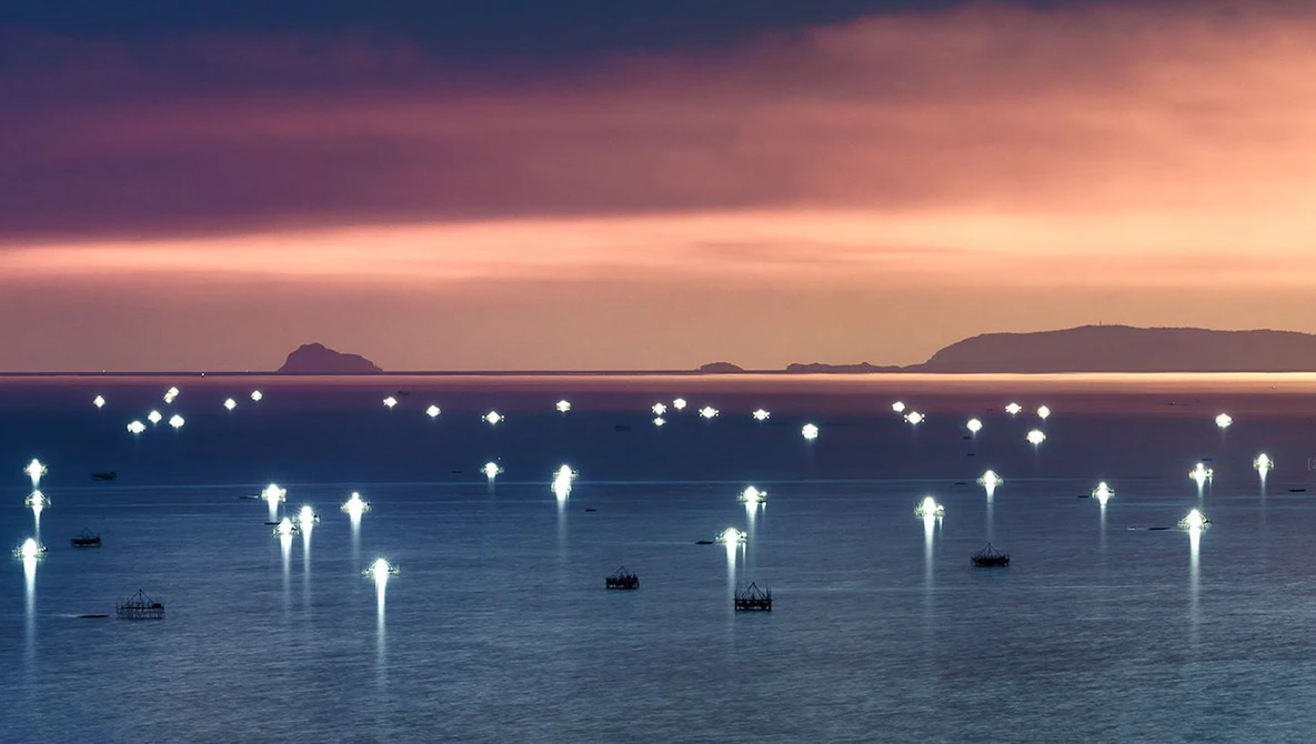 Moored boats with illuminated buoys in calm water at twilight, mountains silhouetted against a purple and pink sky.