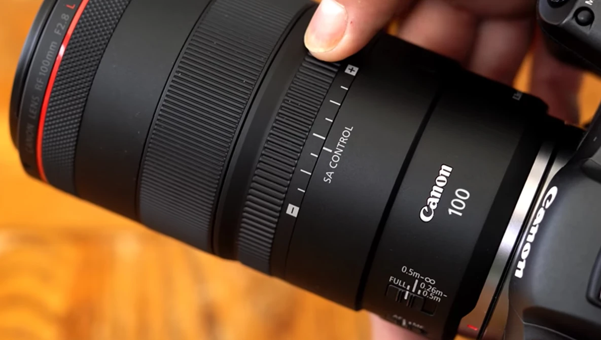 Canon RF 100mm macro lens held in hand against warm orange background.
