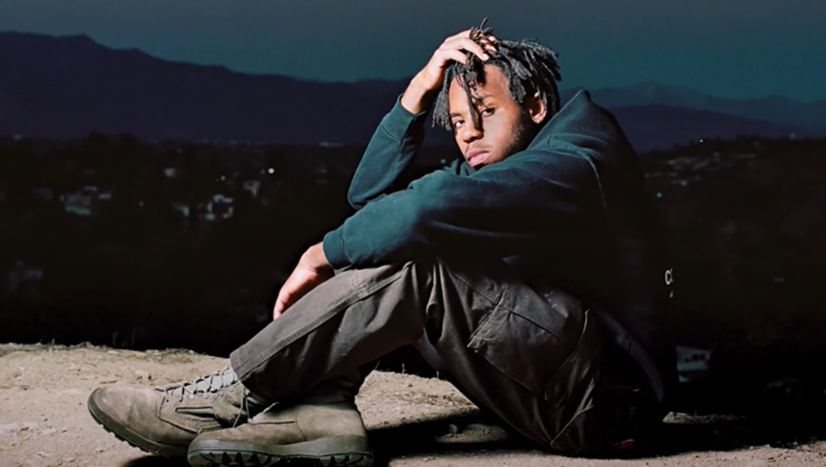 Young man sitting on ground at dusk with head resting on arm, wearing teal jacket and dark pants.