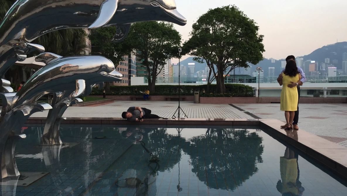 Photographer lying on wet pavement to shoot reflections in still water, with woman in yellow dress posing nearby.