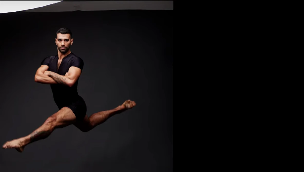 Male dancer in black outfit performing a horizontal leap against dark background.