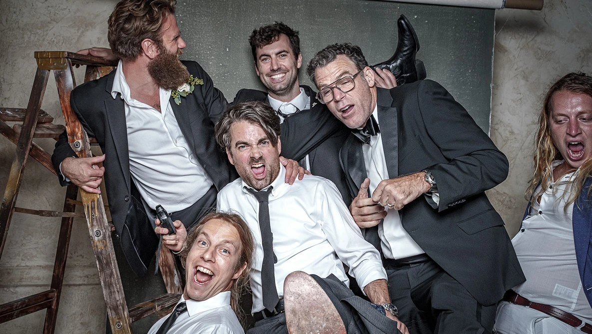 Group of six men in formal attire celebrating exuberantly at an indoor event.