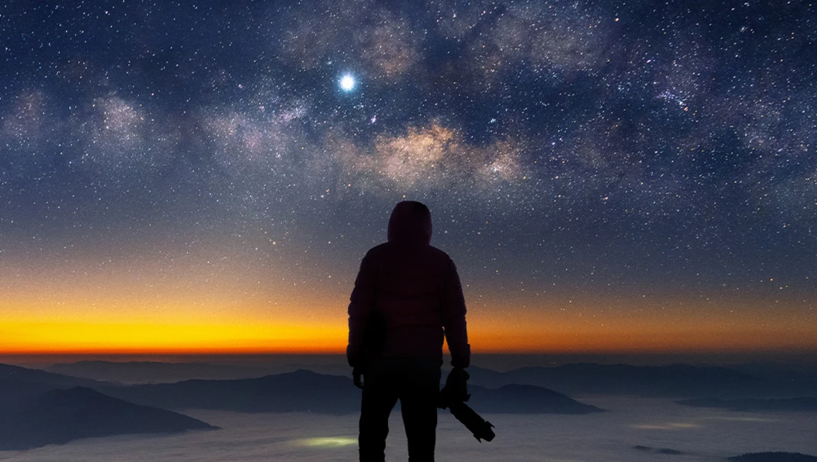 Silhouetted figure standing on a mountain overlooking clouds under a starry night sky with the Milky Way visible.