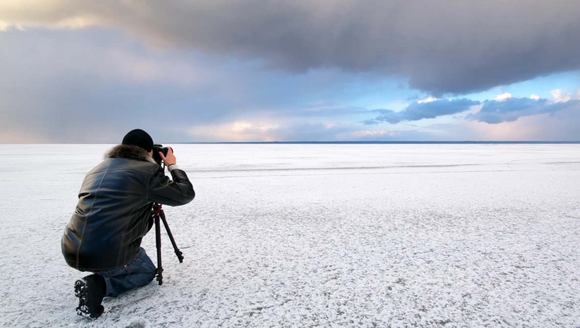 Photographer crouching on a beach with a camera on tripod, framing a seascape with dramatic cloudy sky.