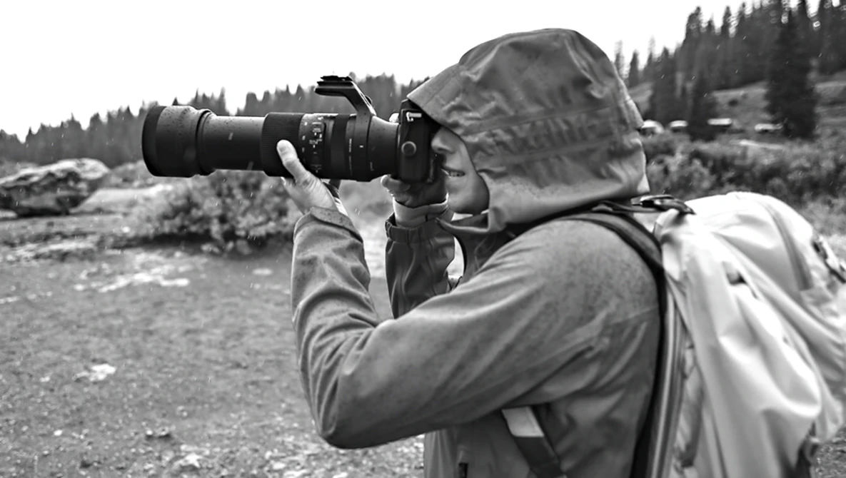 Photographer in hooded jacket shooting with telephoto lens in mountainous terrain.