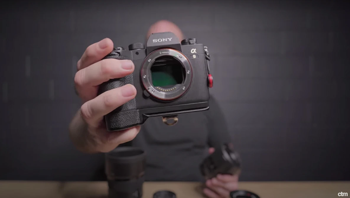 Sony mirrorless camera held in hand, showing open sensor and mount with a black battery grip attached.
