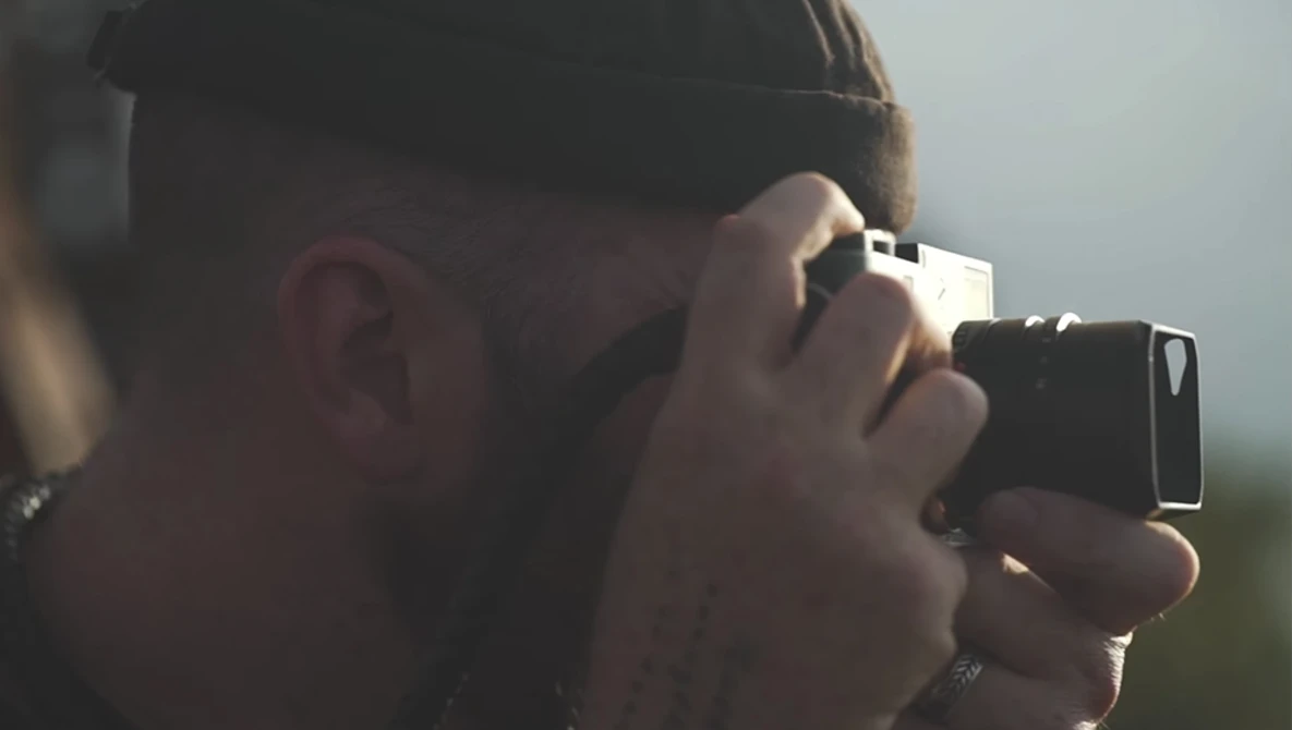 Photographer holding a vintage film camera to their face while wearing a cap and sunglasses.