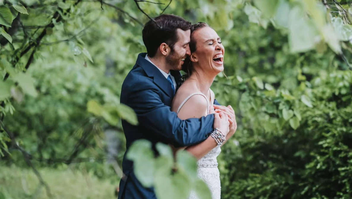 Groom in navy suit embracing laughing bride in white lace dress surrounded by green foliage.