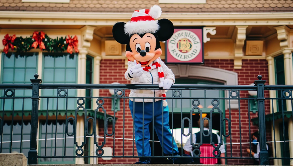 Mickey Mouse character in red and white Santa hat on a balcony railing during holiday decorations.