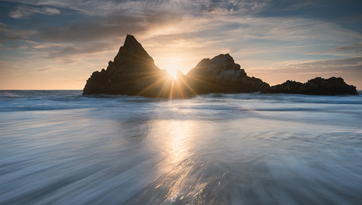 Two rock formations silhouetted against sun rising between them in ocean during golden hour.