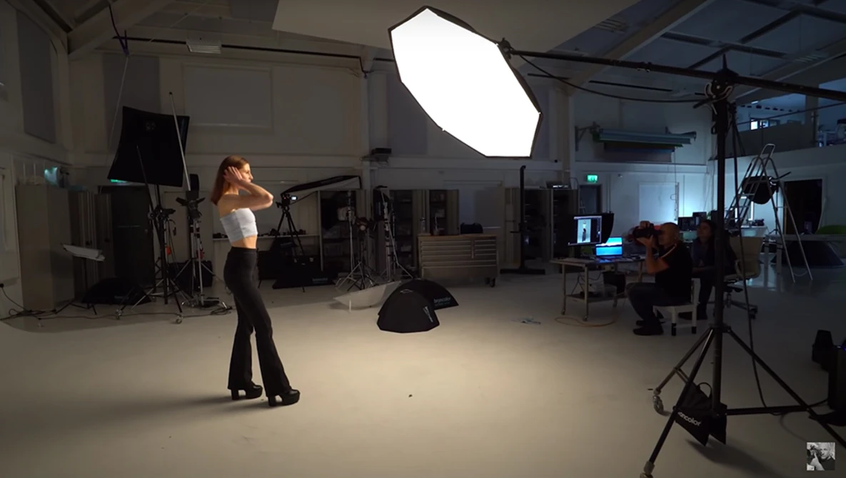 Female model posing during a studio photoshoot with professional lighting setup and crew members present.