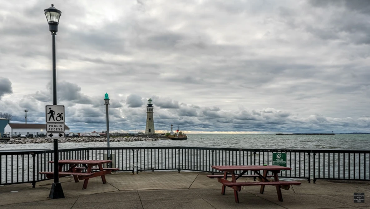 Waterfront pier with picnic tables, lamp post, and lighthouse visible across the water under dramatic cloudy sky.
