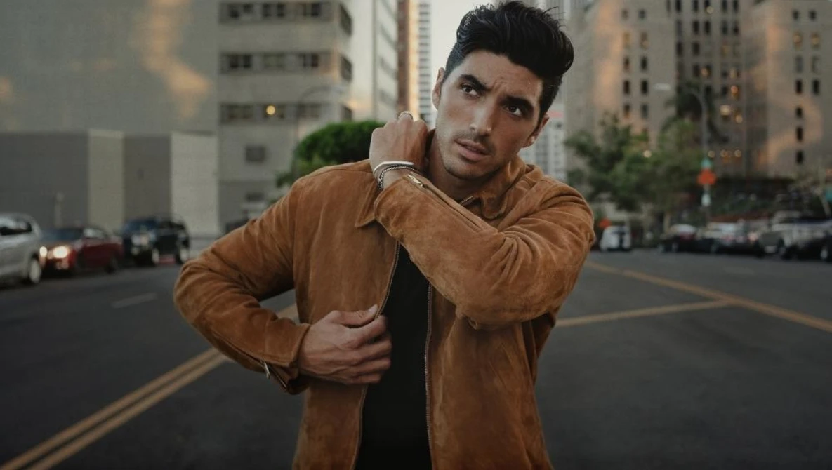 Man in tan suede jacket posing on urban street with city buildings in background.