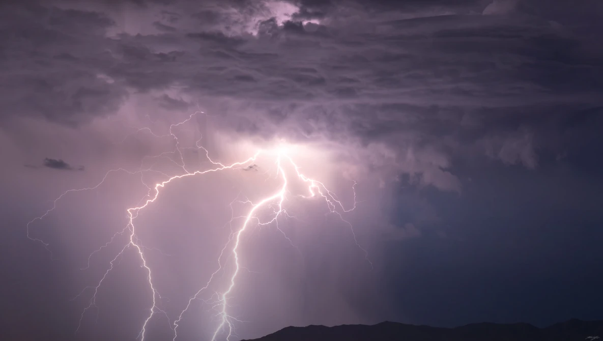 Multiple lightning bolts strike from storm clouds illuminated by bright white light against a dark purple sky.