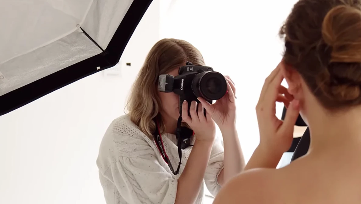 Photographer holding a film camera to her face while being photographed by another person in a studio setup.