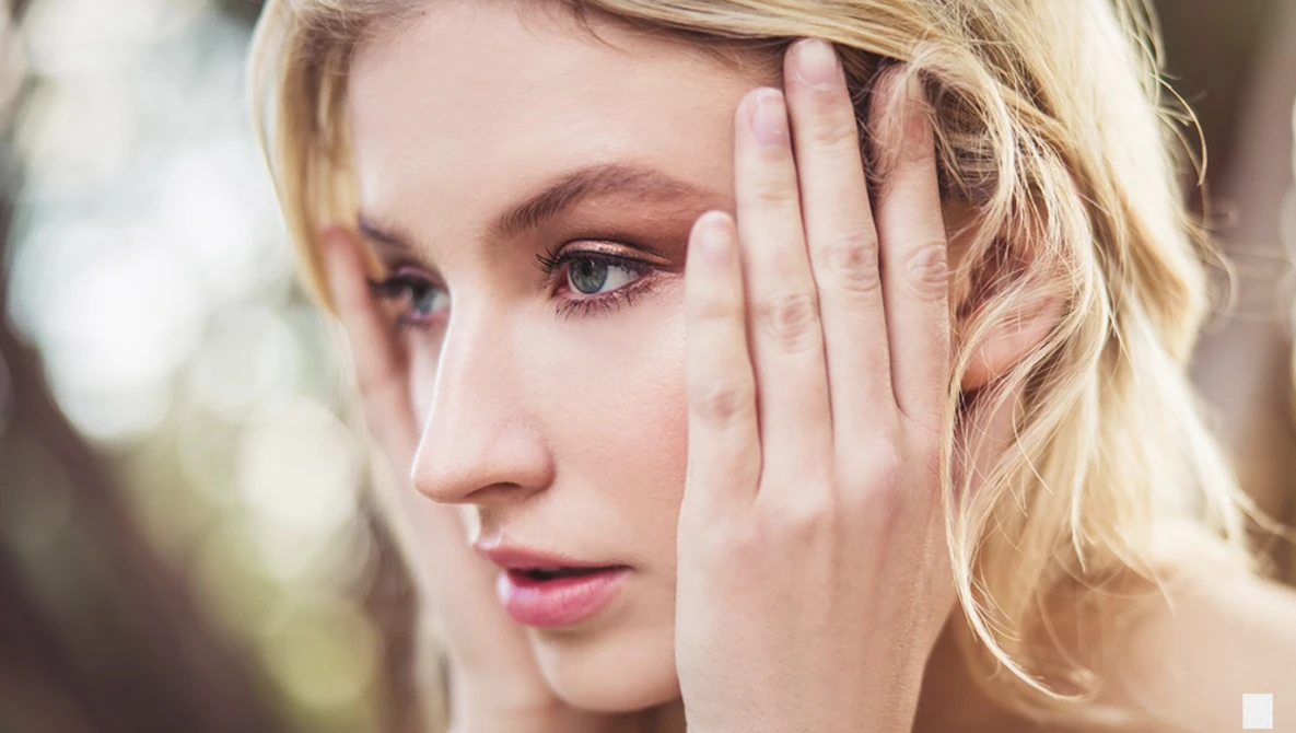 Blonde woman in profile with hand near face, soft natural lighting and blurred background.