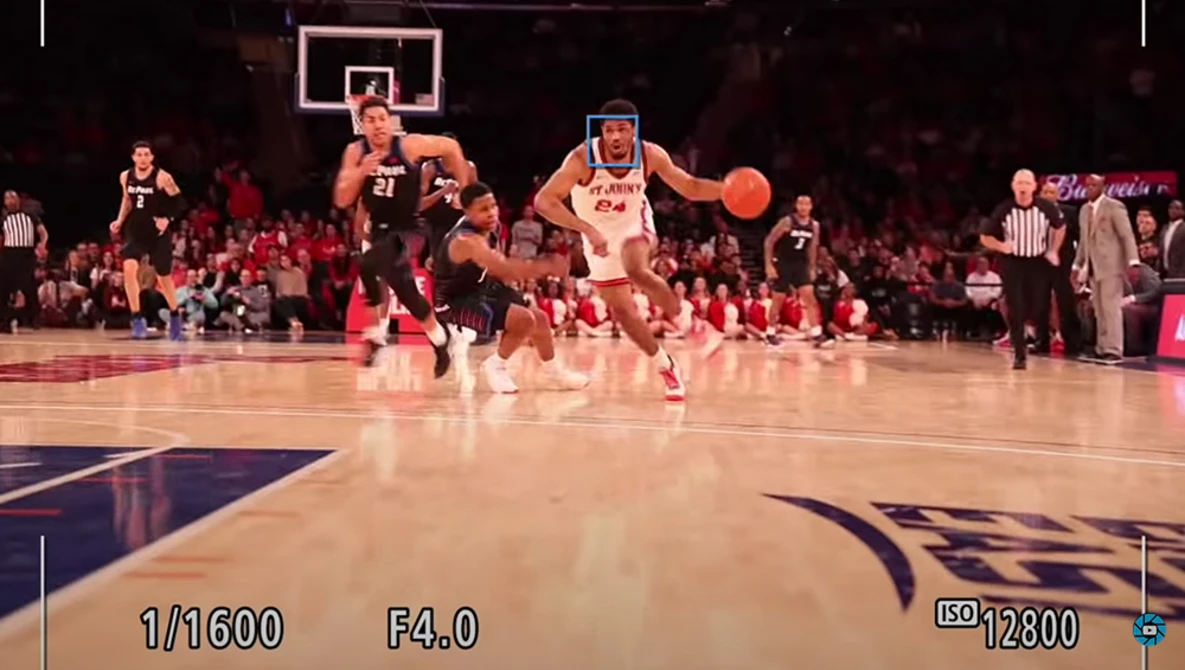 Basketball player in white jersey dribbling ball during live game with packed arena in background.