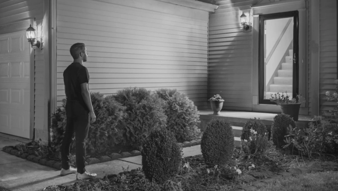 Man in dark clothing standing on a residential porch at night, surrounded by landscaped shrubs.