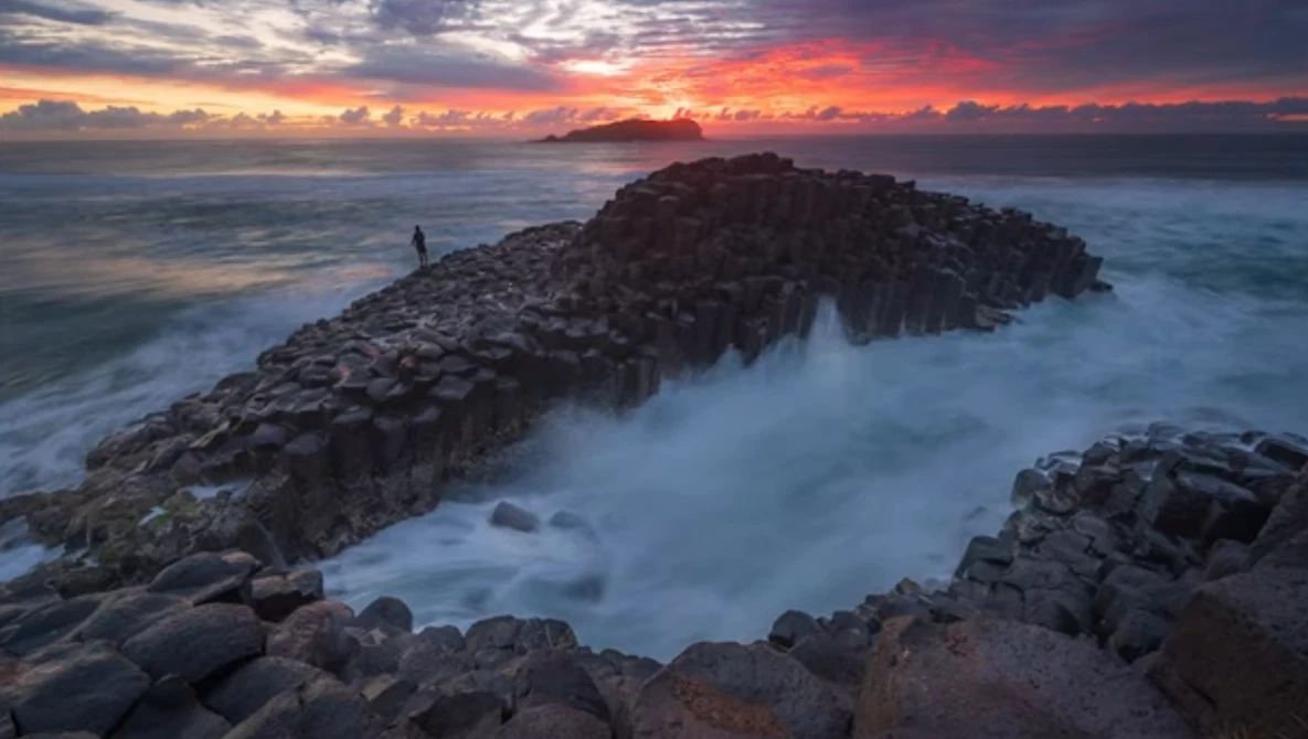 Rocky coastal headland with a solitary figure silhouetted against a dramatic sunset sky over the ocean.
