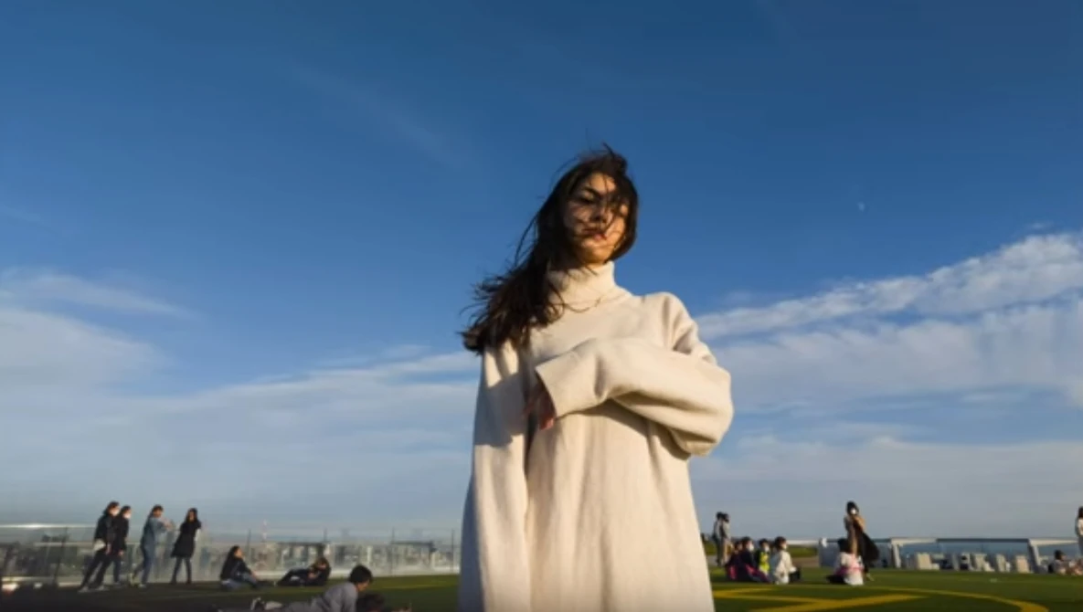 Woman in cream sweater standing outdoors against blue sky with distant cityscape.