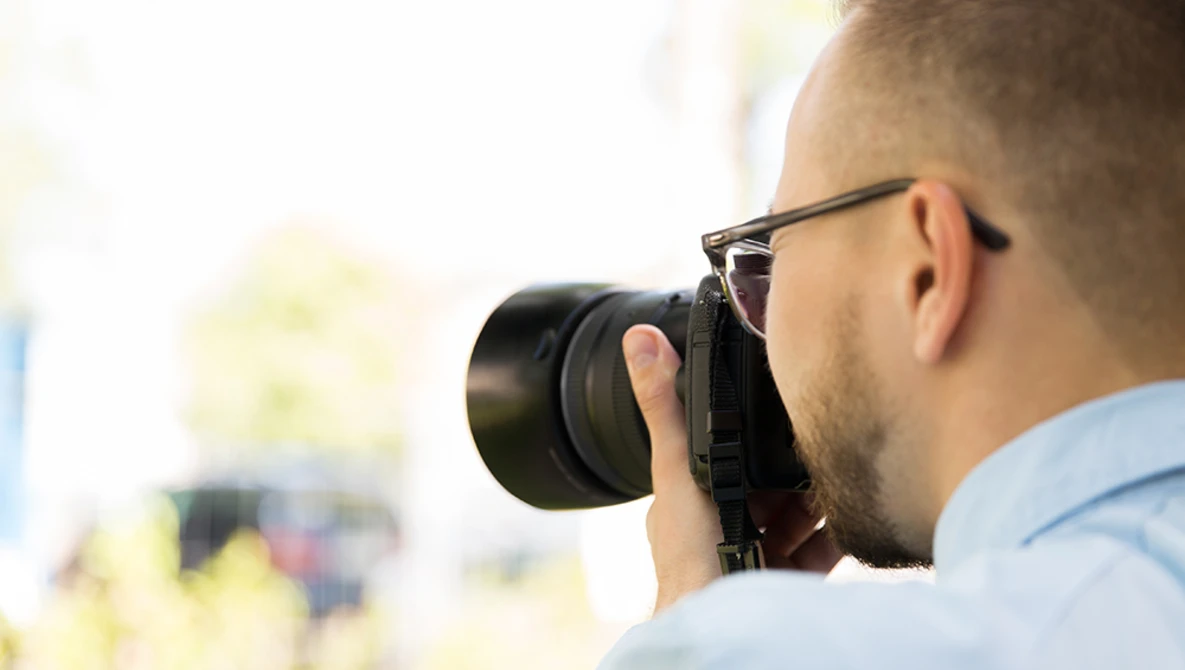 Male photographer wearing glasses and light blue shirt holding camera up to face while shooting outdoors.