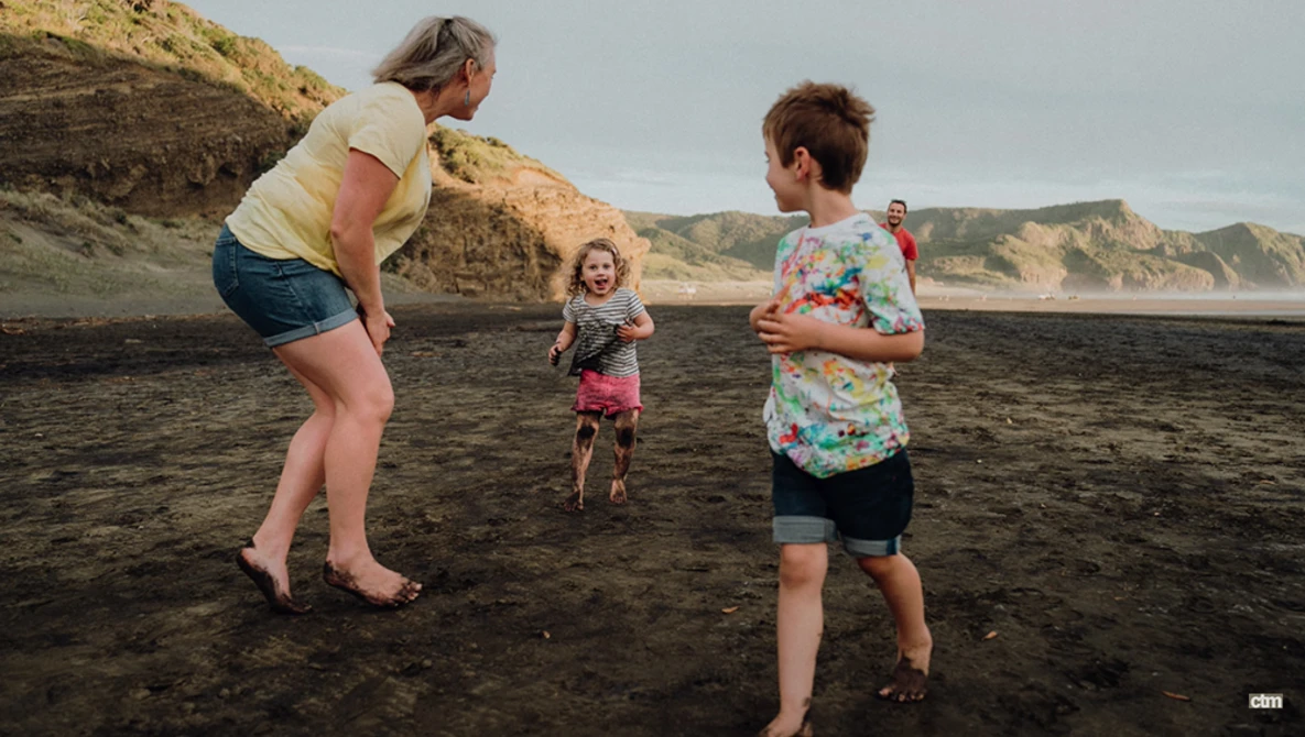 An adult and three children playing together on a dark sand beach backed by tall cliffs.
