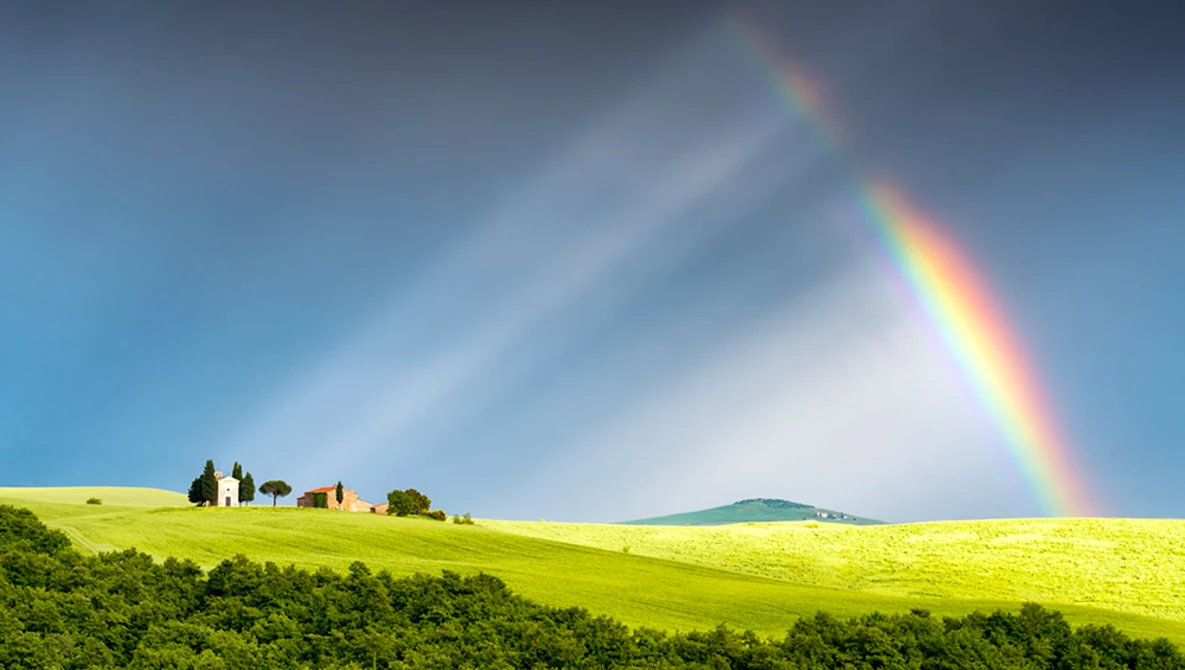 Double rainbow arcing over a green hillside with a farmhouse and cypress trees beneath stormy skies.
