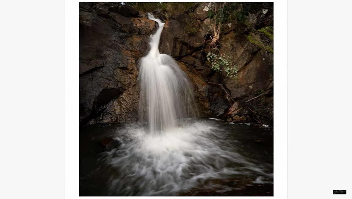 Small waterfall cascading into a rocky pool surrounded by dark stone walls and moss-covered vegetation.