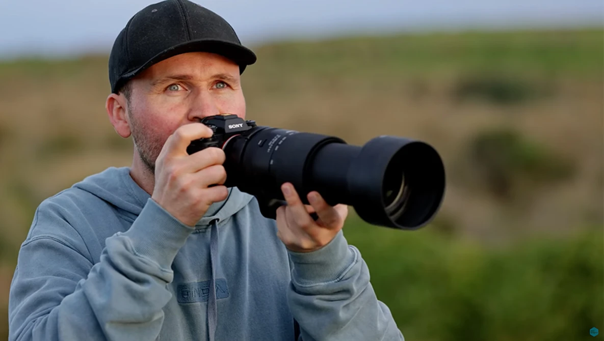 Photographer holding a telephoto lens while looking through the viewfinder in an open field.