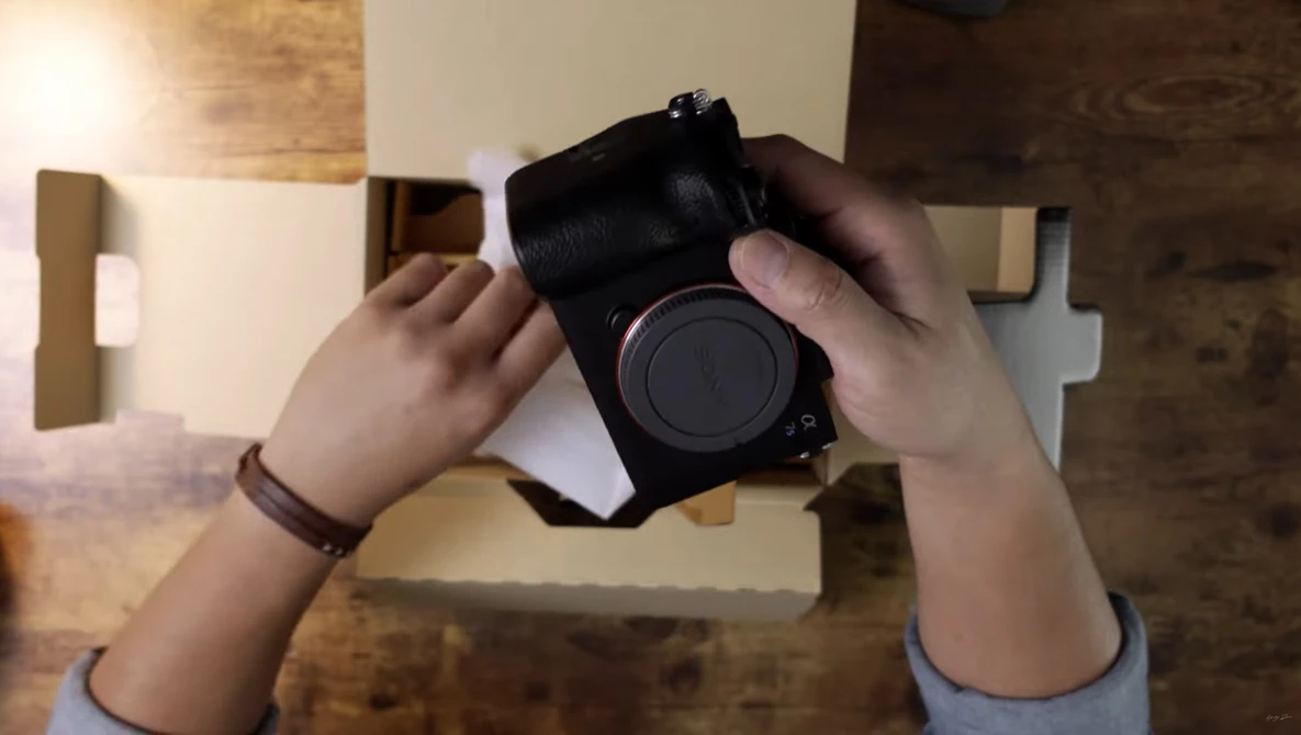 Hands holding a mirrorless camera body with lens cap removed, photographed from above on a wooden surface.