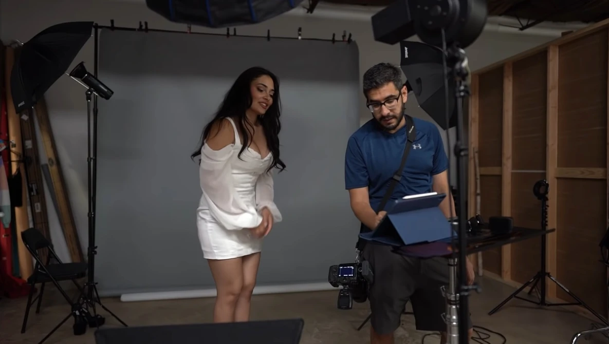 Photographer reviewing footage on tablet while model poses in white dress during studio shoot.