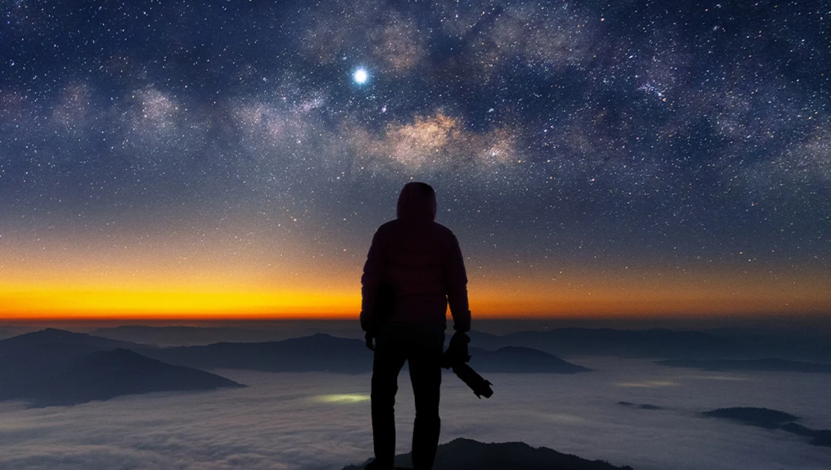 Silhouetted figure standing on a mountain peak gazing at the Milky Way and sunrise.